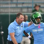 Football coach instructing player wearing green helmet and blue Roll Wave shirt during practice session