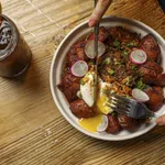 Bowl of fried plantains with poached egg, shredded meat, and radish slices on a wooden table with iced tea.