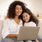 a person and a girl sitting on a couch looking at a laptop