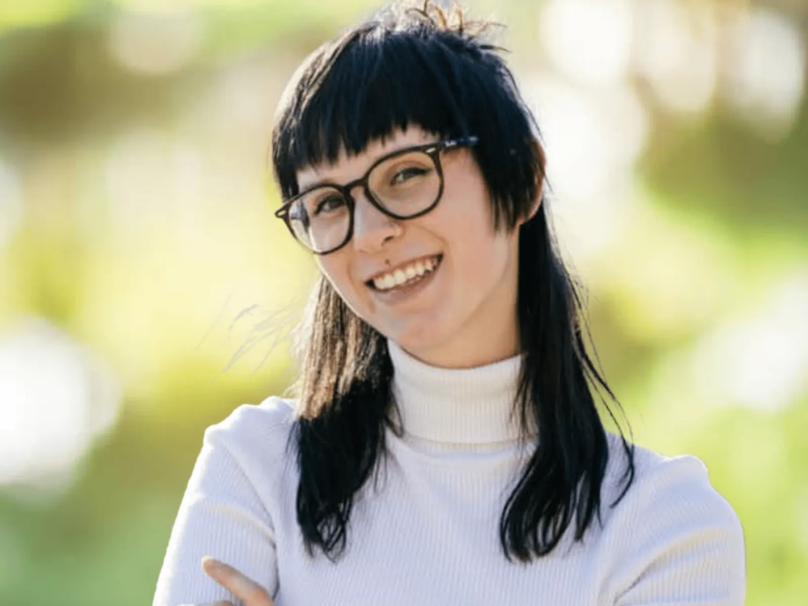 Smiling young woman with glasses wearing a white turtleneck in a bright outdoor setting with blurred greenery.