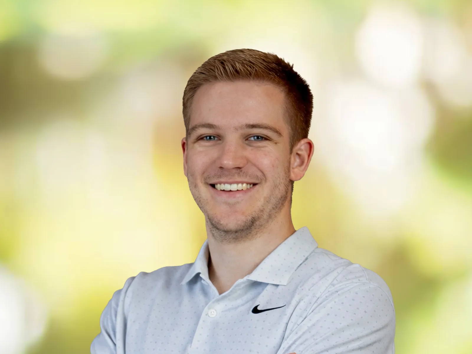 Smiling young man with short blond hair wearing a white polo shirt against a blurred green background