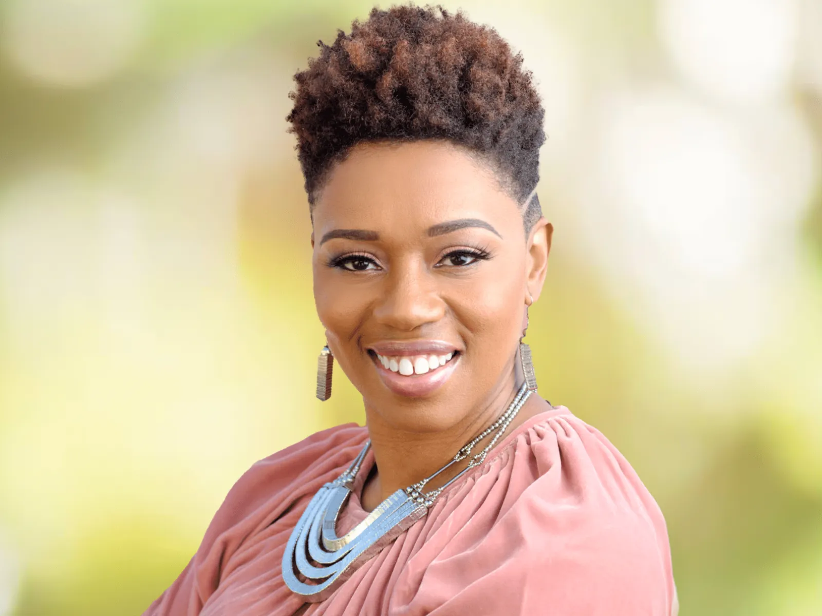 Confident woman with short curly hair in pink blouse and silver necklace smiling against soft blurred background