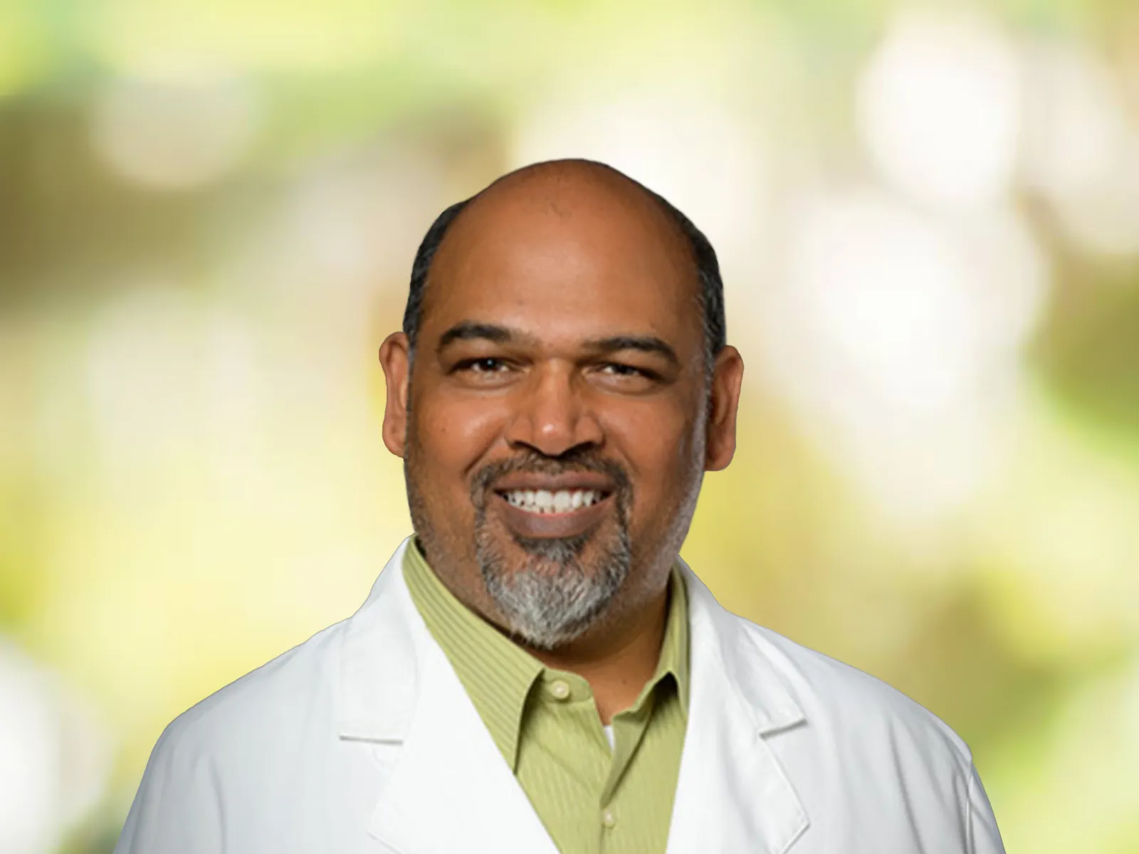Smiling male doctor in white coat with Dr. Rai embroidered, standing against a soft green blurred background.