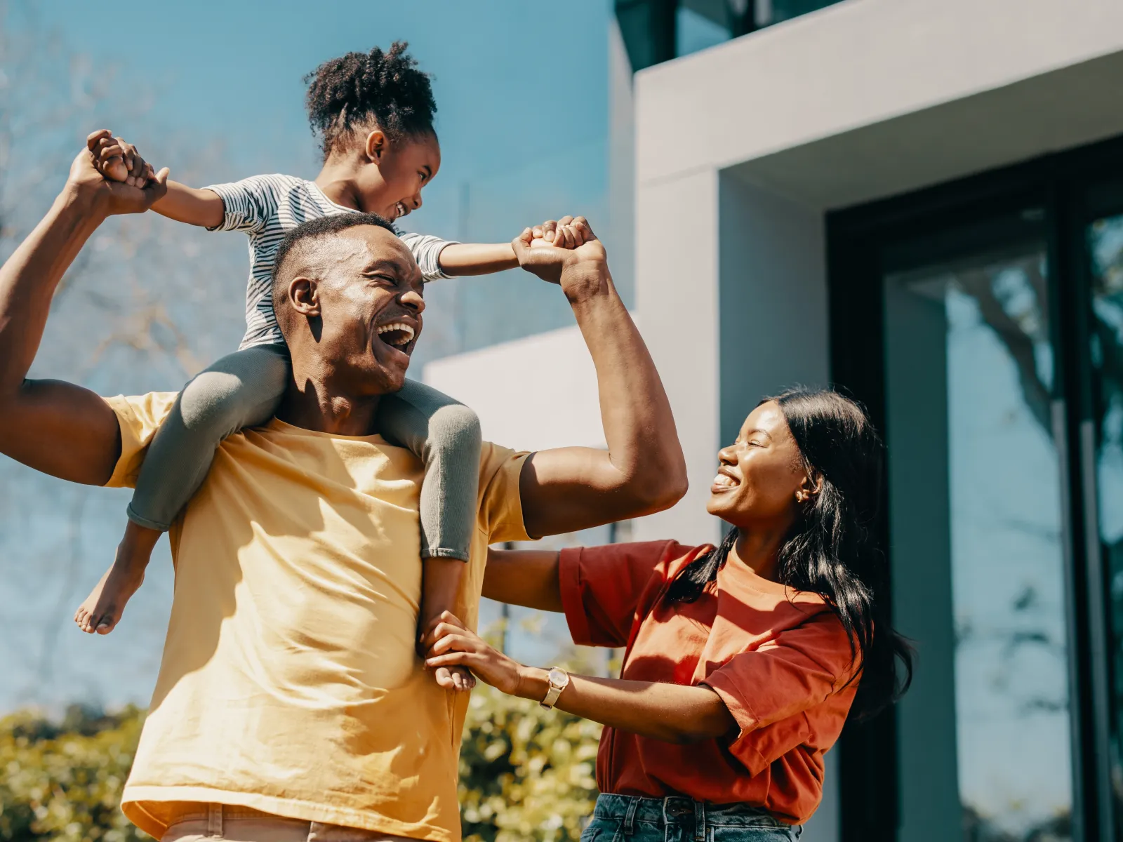 Happy family outdoors with father carrying daughter on shoulders and mother smiling nearby in sunny day.