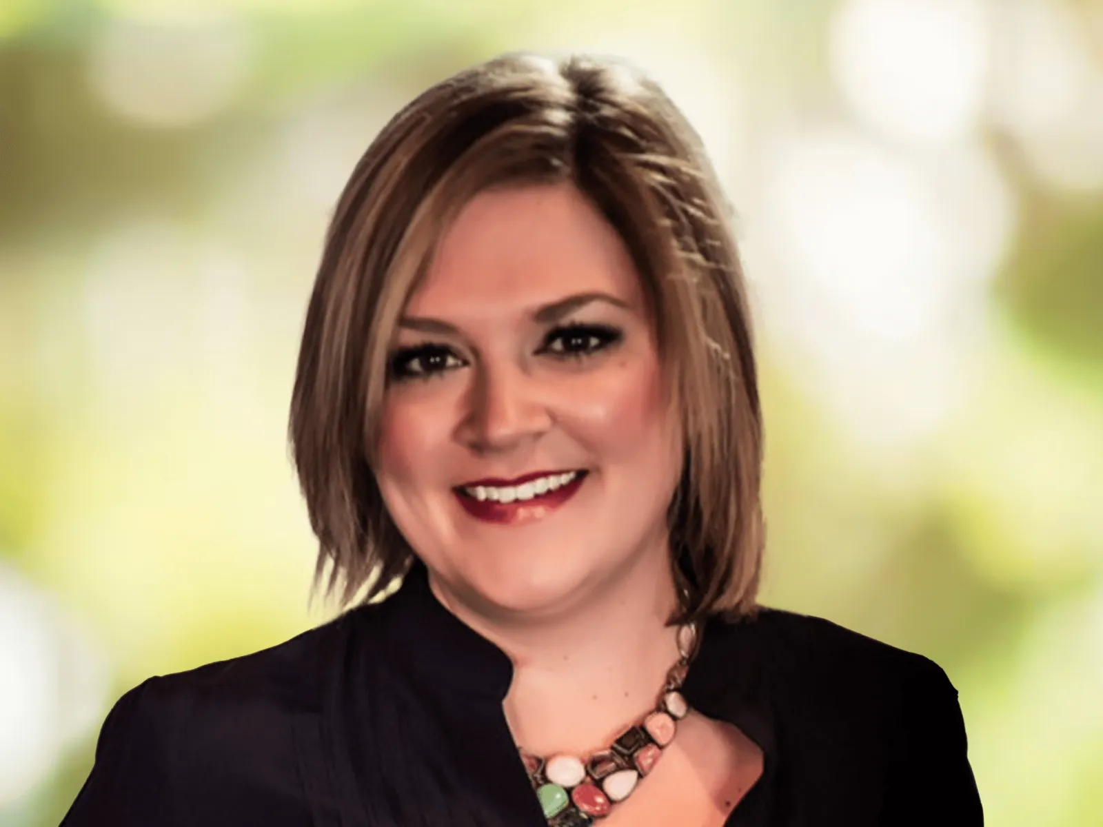 Smiling woman with short blonde hair in a black top and colorful beaded necklace against a blurred green background