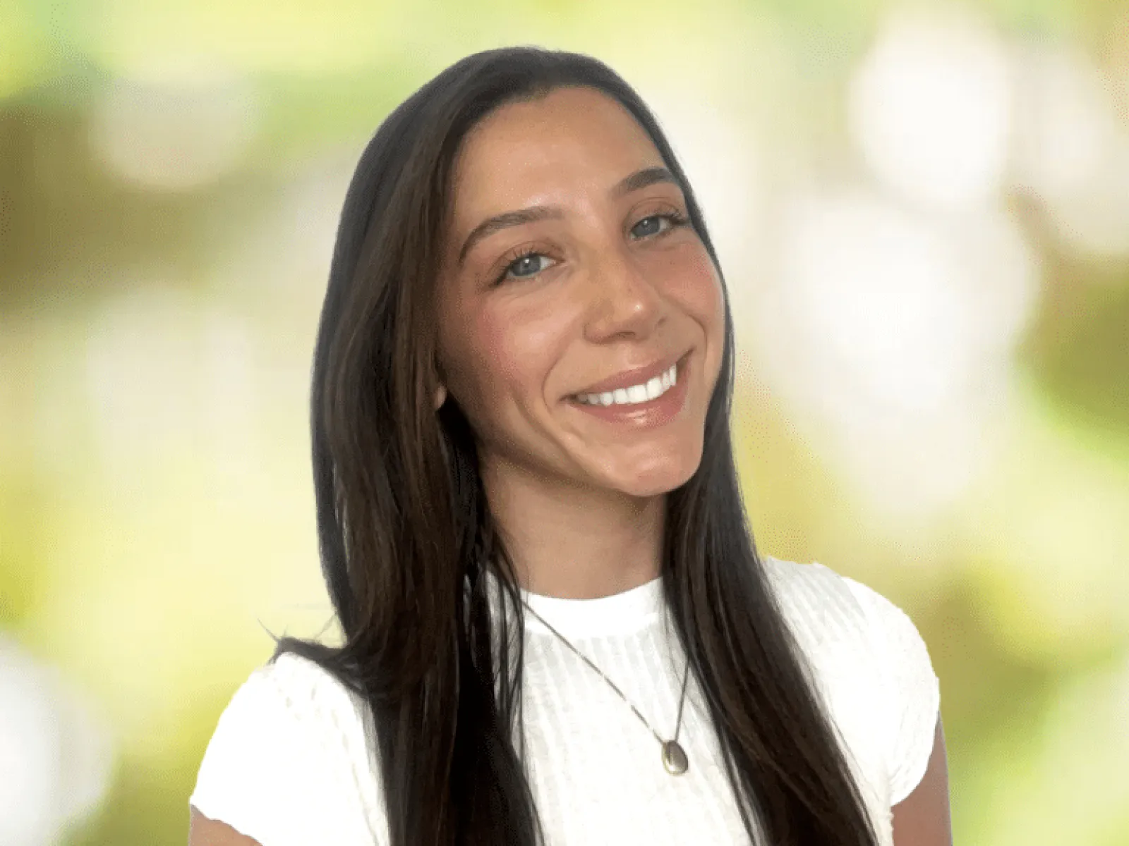 Smiling woman with long dark hair wearing a white top and pendant necklace against a blurred nature background