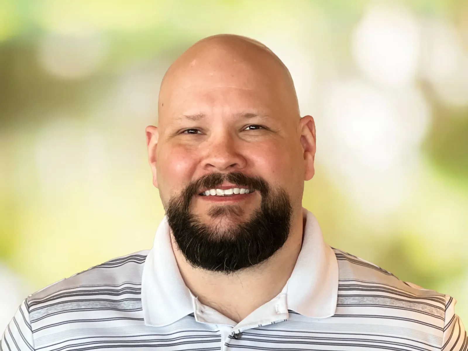 Smiling bald man with beard wearing a white and gray striped polo shirt against a blurred green background