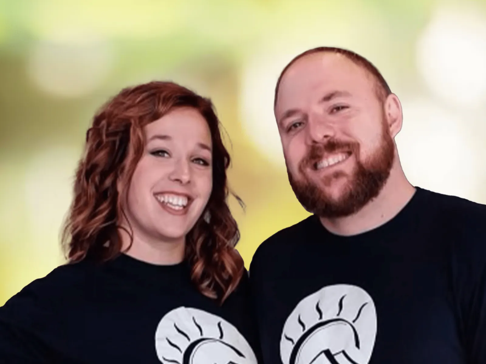 Smiling man and woman wearing matching black t-shirts with mountain and sun logo on blurred green background