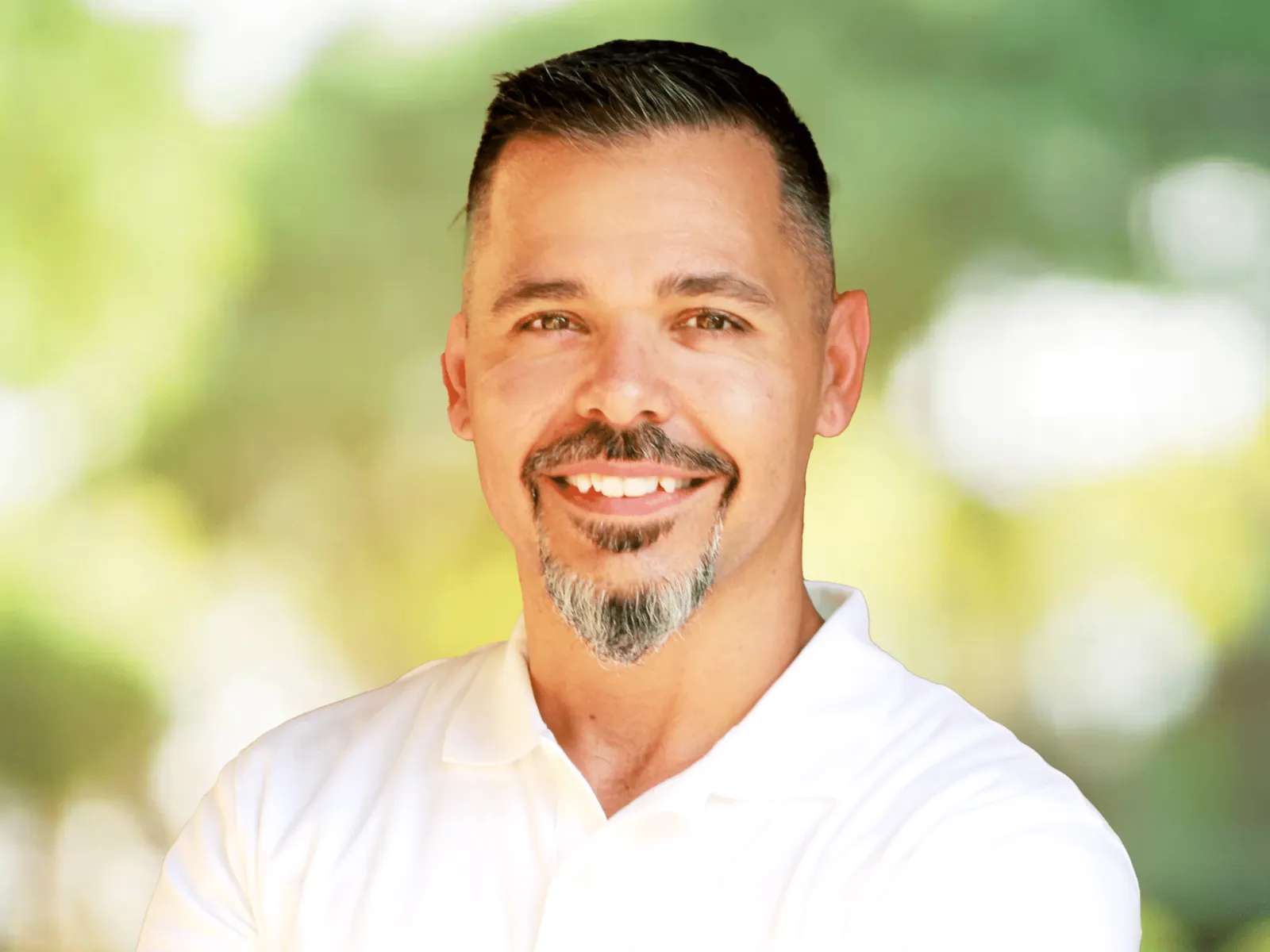 Smiling man with short black hair and goatee wearing a white polo shirt outdoors with blurred green background