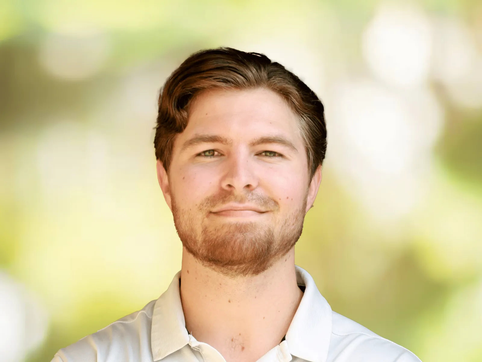 Smiling man in white and gray polo shirt with 100% chiropractic logo against a blurred green background.