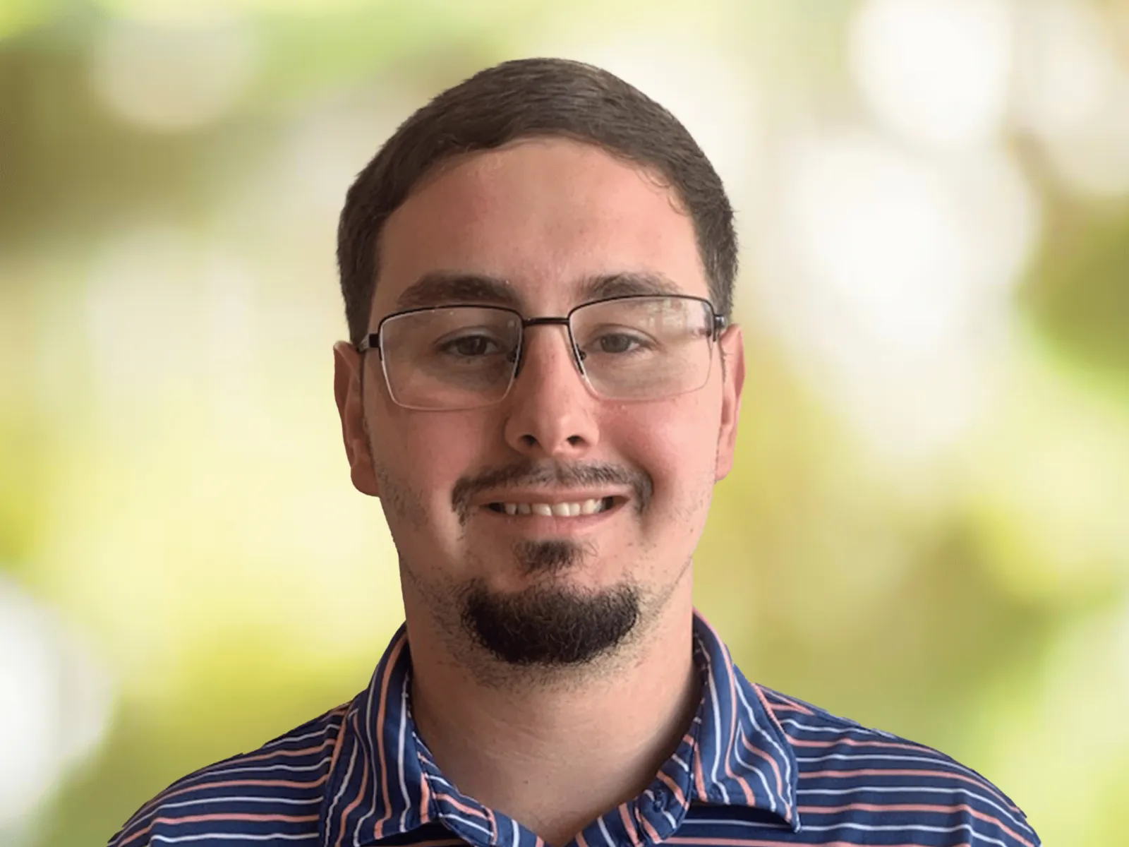 Young man with glasses and beard smiling wearing a blue striped collared shirt against blurred green background