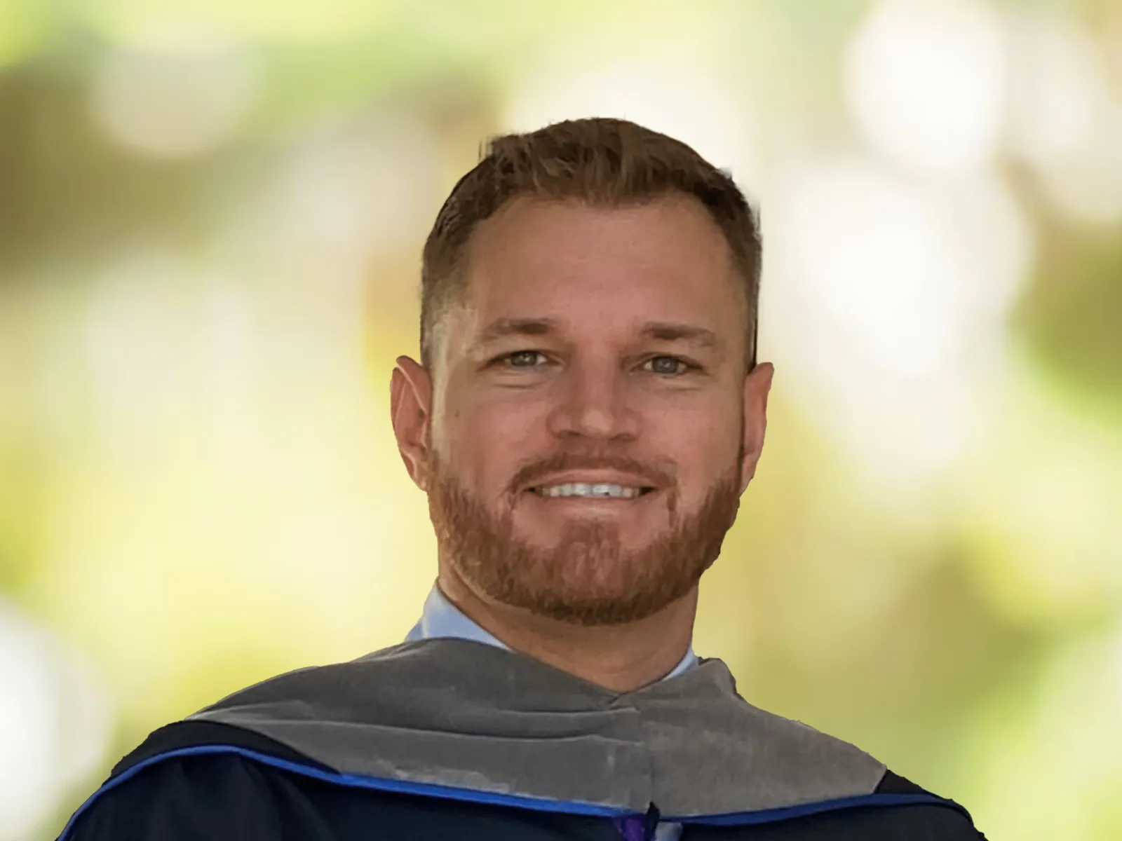 Smiling man in graduation gown with blurred green and yellow outdoor background