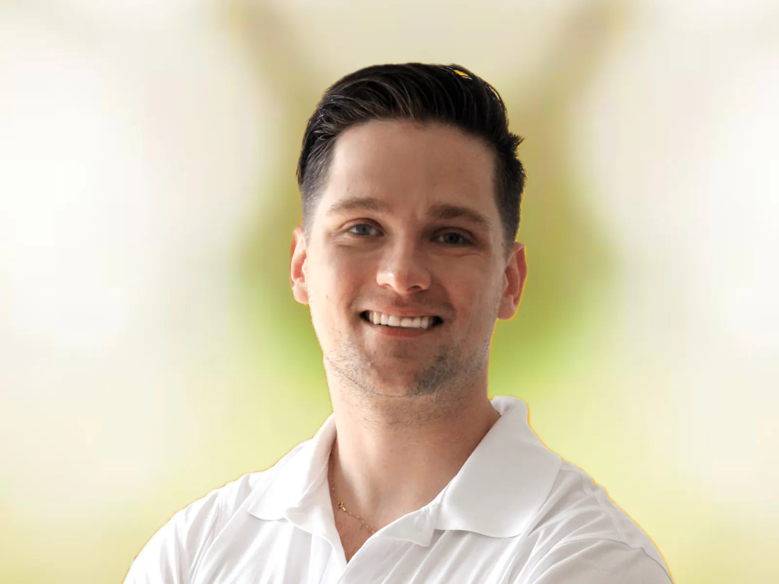 Smiling man in white polo shirt with 100% chiropractic logo stands against blurred green background.