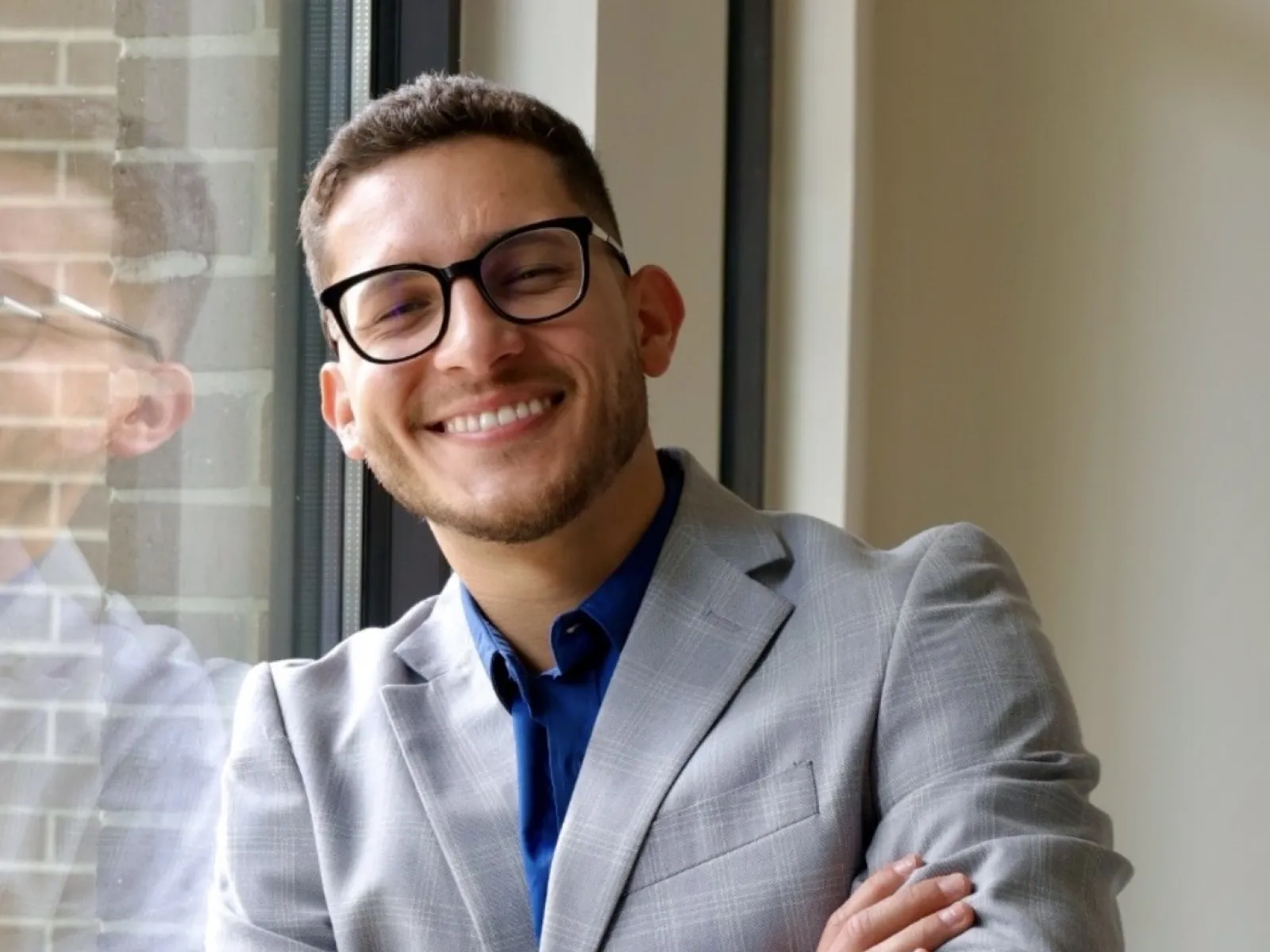 Smiling young man in gray suit and blue shirt standing with arms crossed by a window indoors.