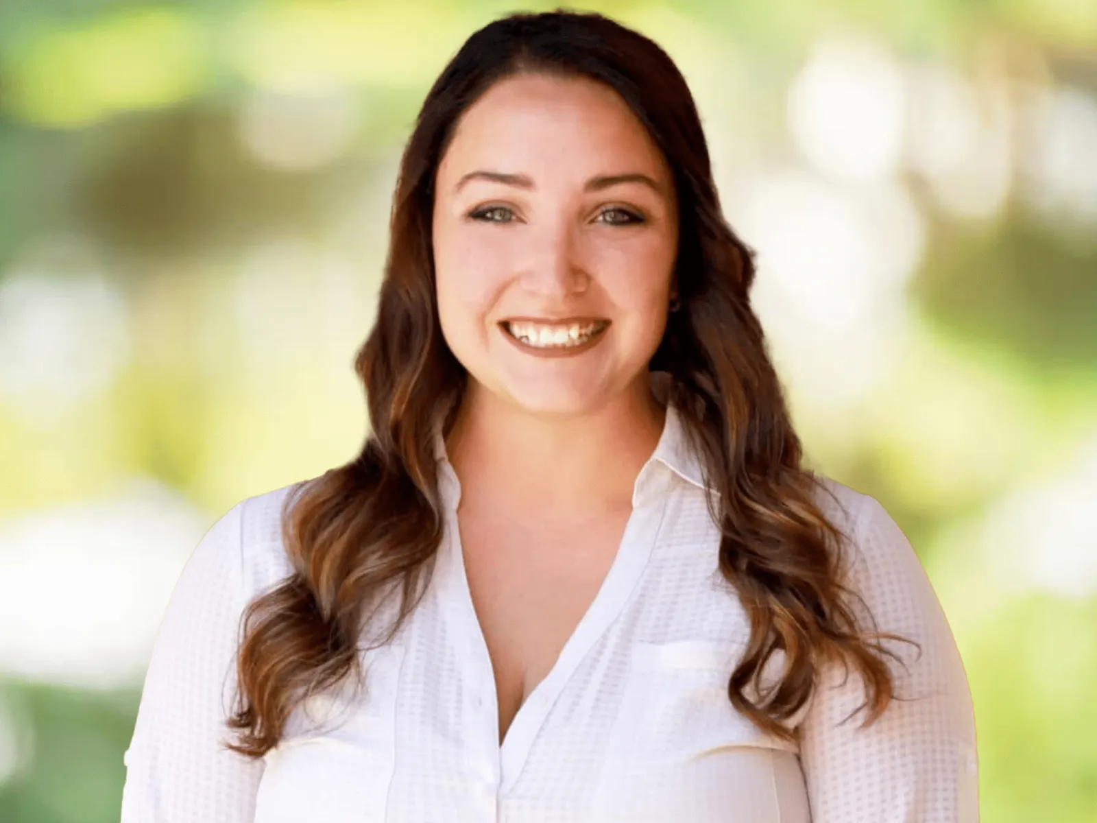 Smiling woman with long brown hair wearing a white button-up shirt in soft outdoor lighting