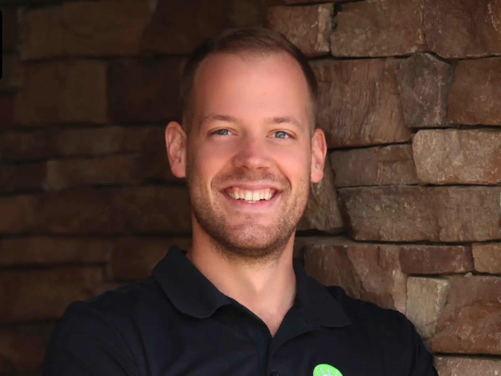 Smiling man in black polo with 100% chiropractic logo stands with arms crossed against stone wall.