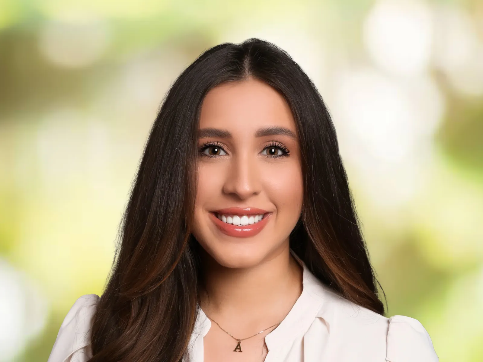Smiling young woman with long dark hair wearing a white blouse and letter A necklace, green blurred background.