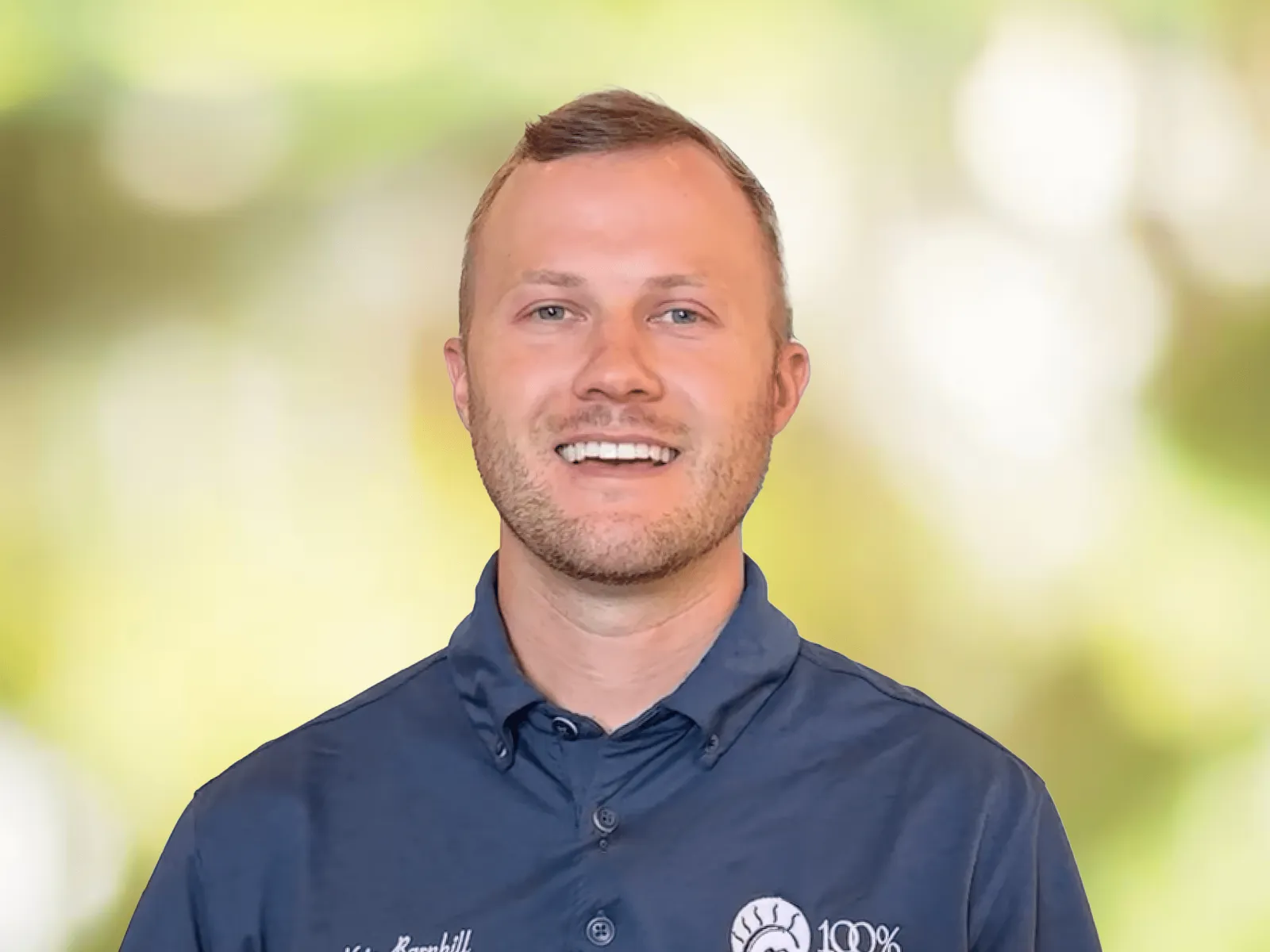 Smiling man in white and gray polo shirt with 100% chiropractic logo against a blurred green background.