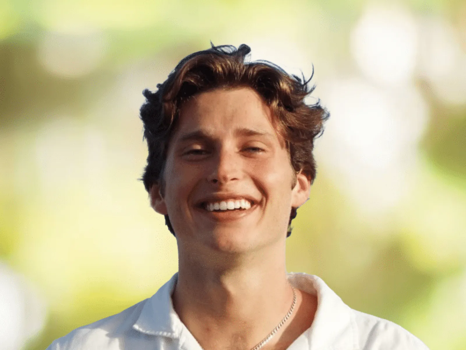 Smiling young man with wavy brown hair wearing a white shirt and silver chain, blurred green outdoor background.