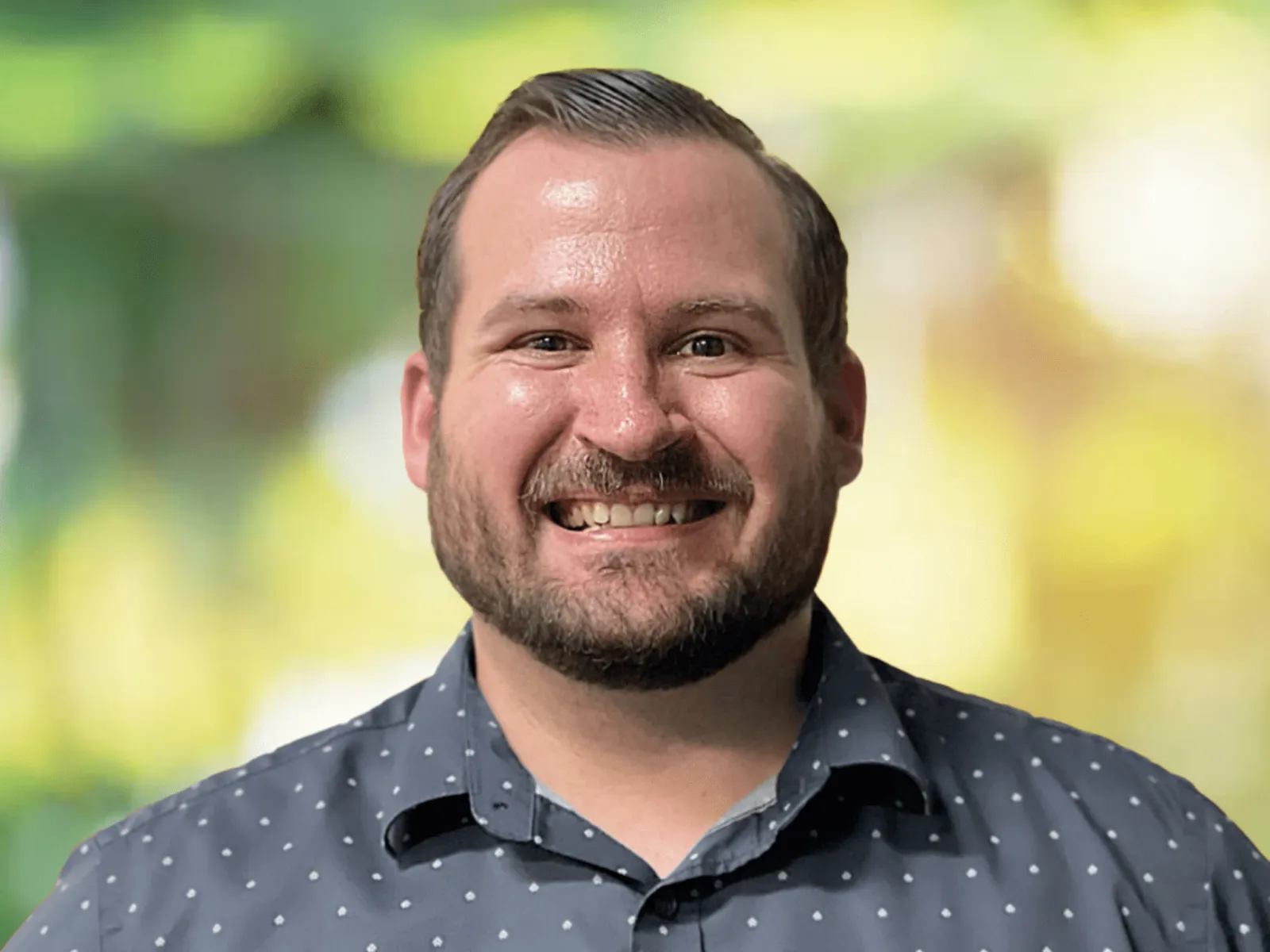 Smiling man with beard wearing a blue patterned shirt with blurred green and yellow background