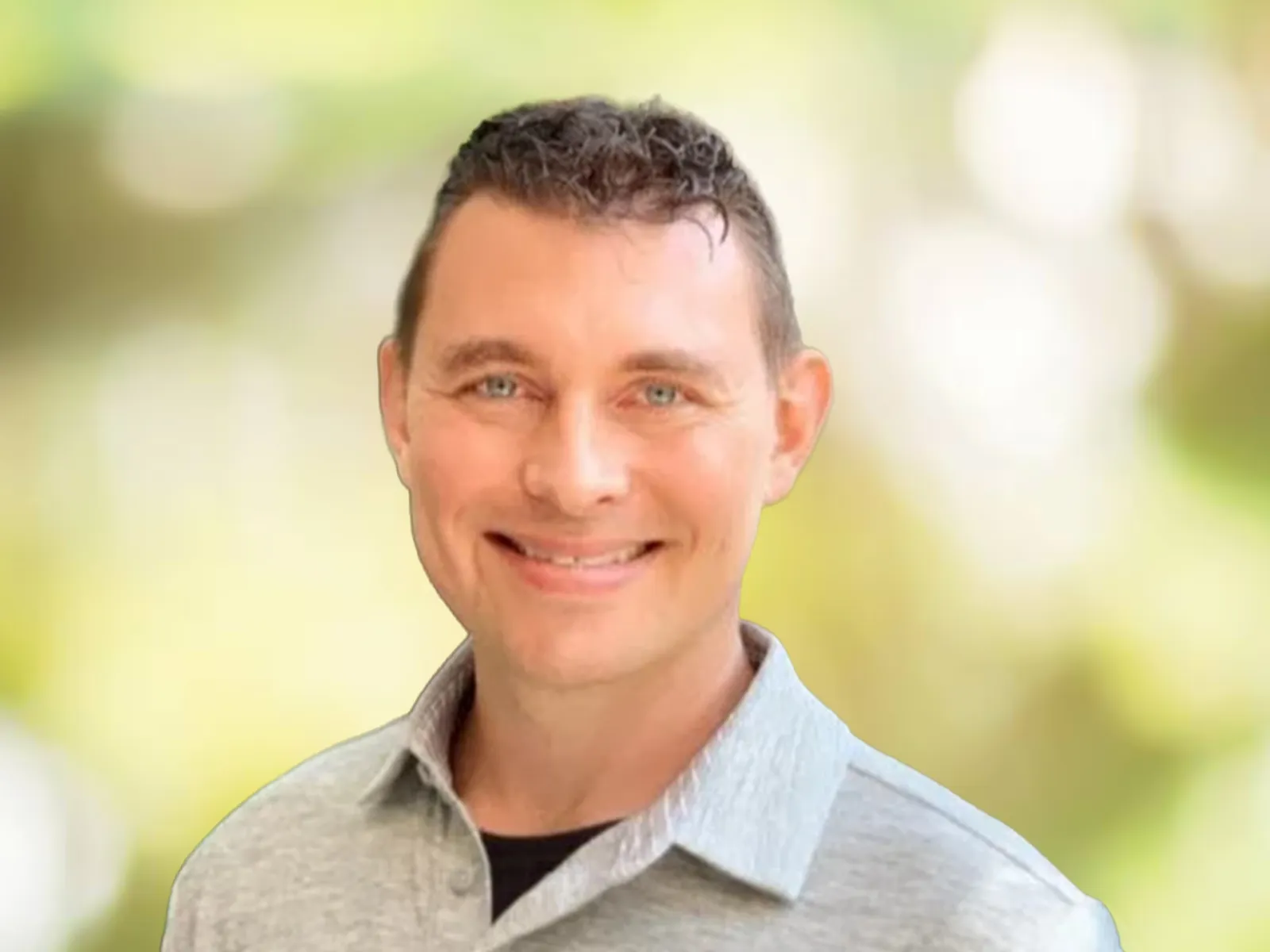 Smiling man with short curly hair wearing a gray collared shirt, blurred green outdoor background.