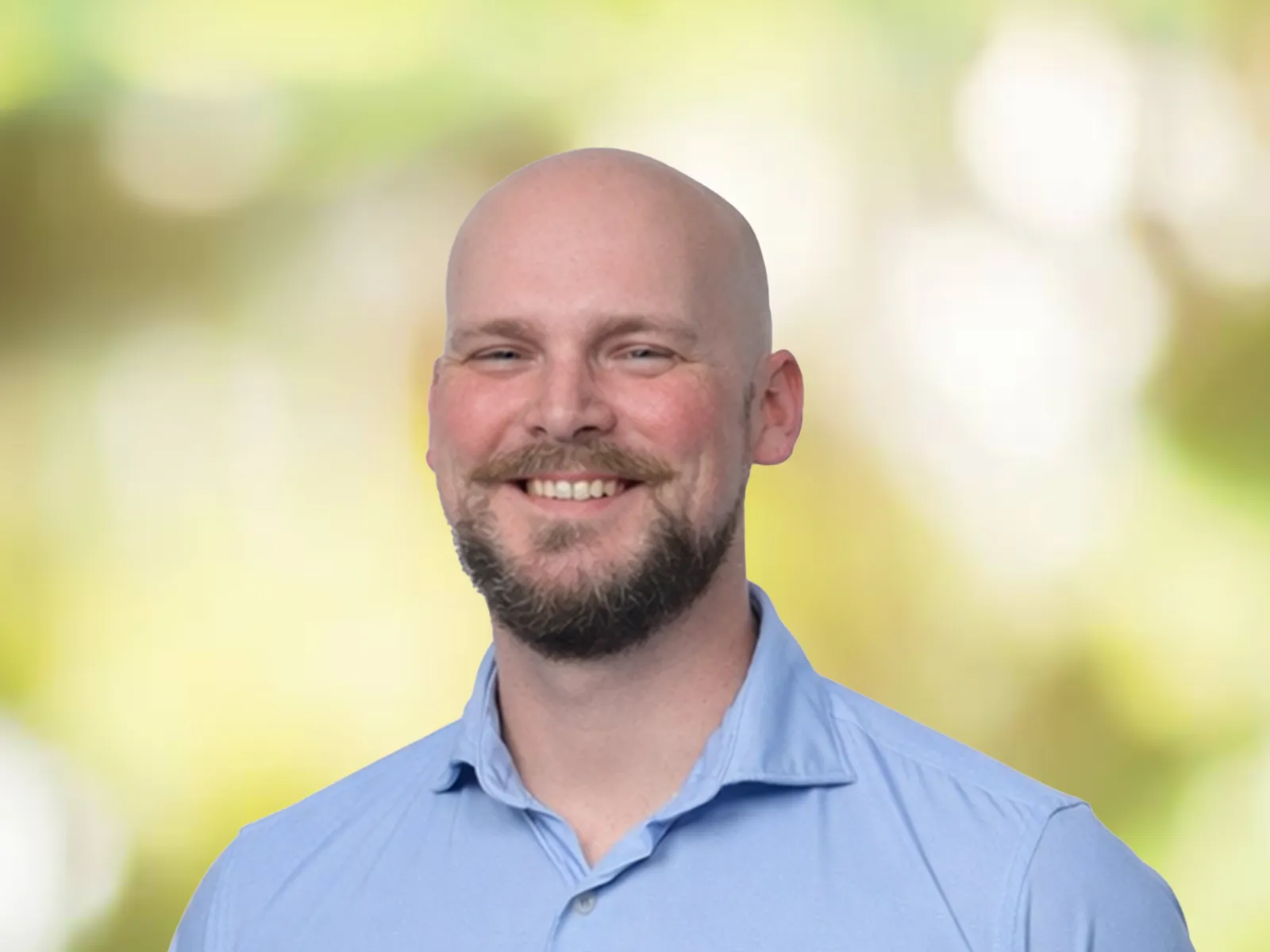Smiling bald man with a beard wearing a light blue shirt against a soft yellow-green blurred background