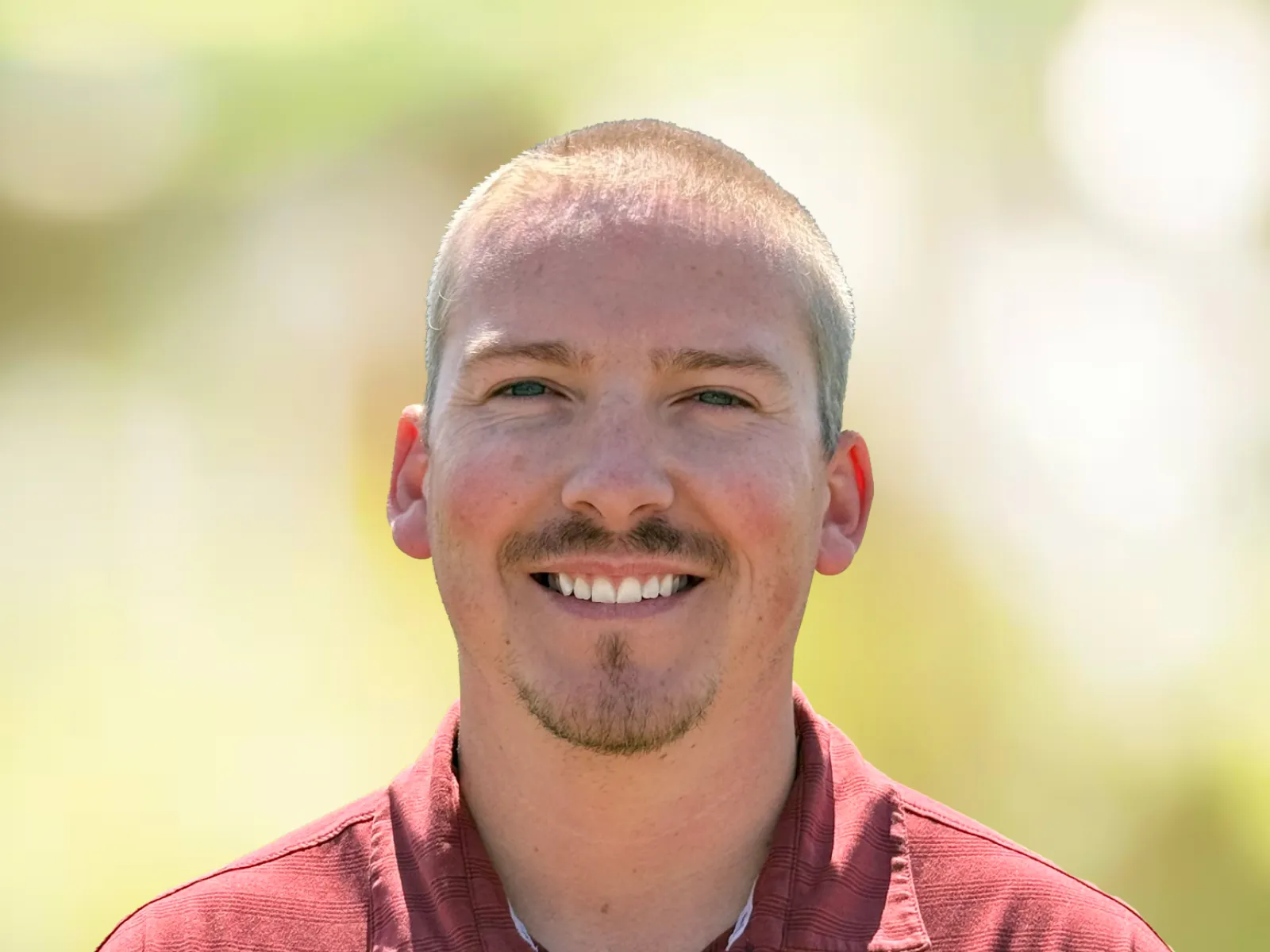 Smiling man in maroon polo shirt with embroidered chiropractic logo and name outdoors with blurred green background