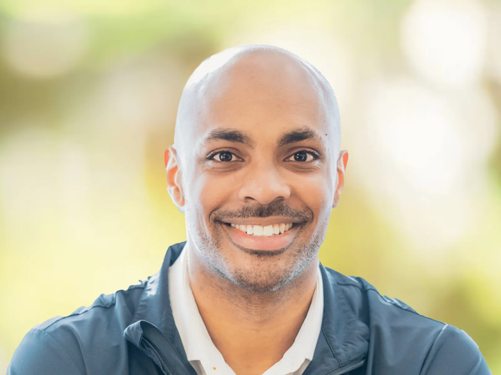 Smiling man wearing a navy jacket with 100% chiropractic logo, blurred green and yellow background