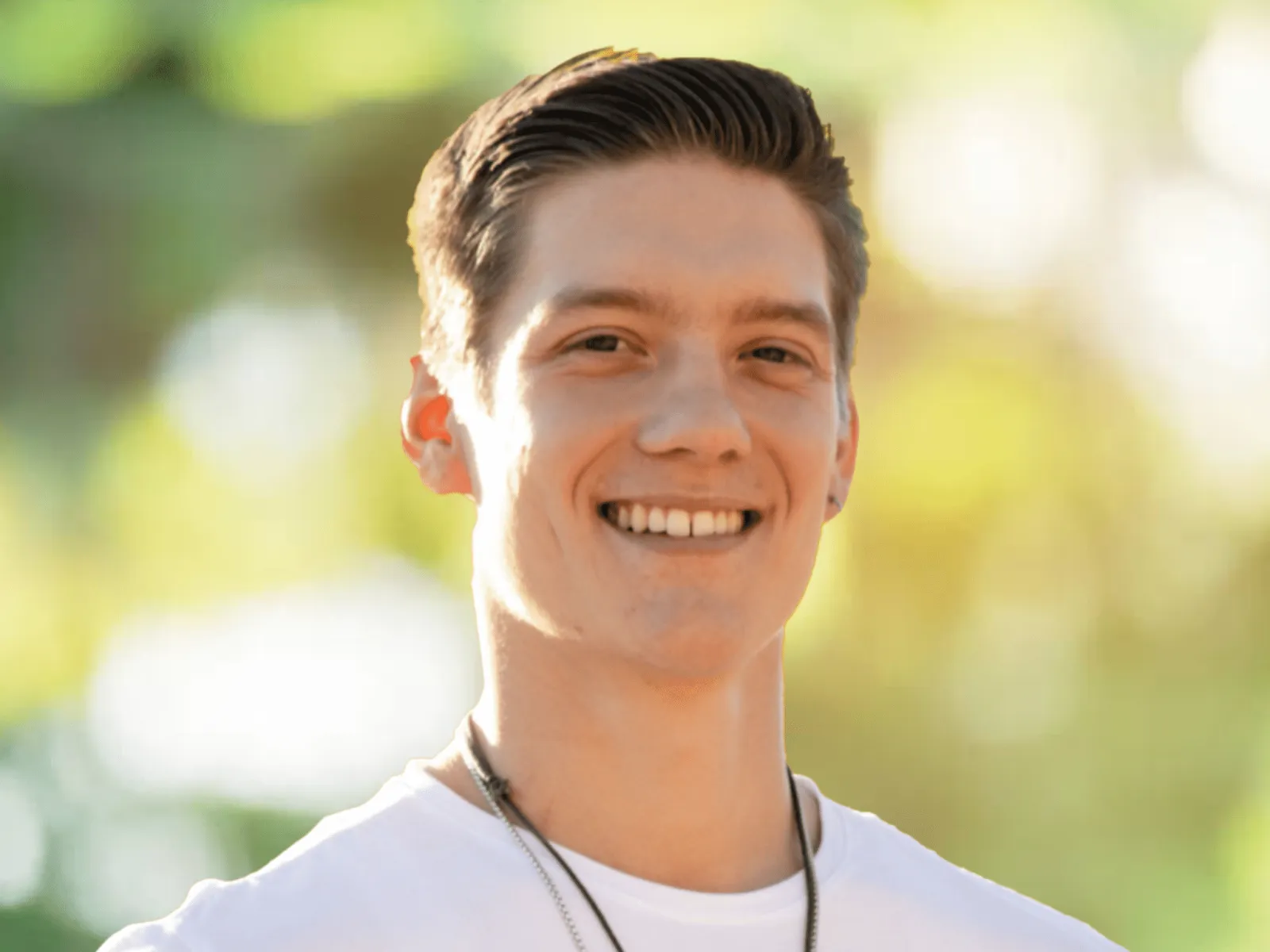 Young man smiling outdoors with sunlight and blurred greenery background wearing white shirt and necklace