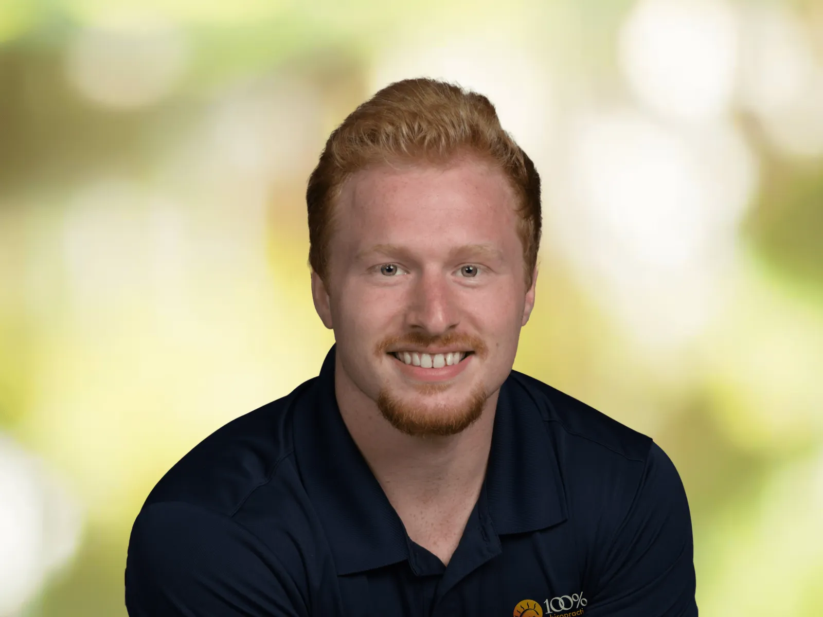Smiling man in white and gray polo shirt with 100% chiropractic logo against a blurred green background.