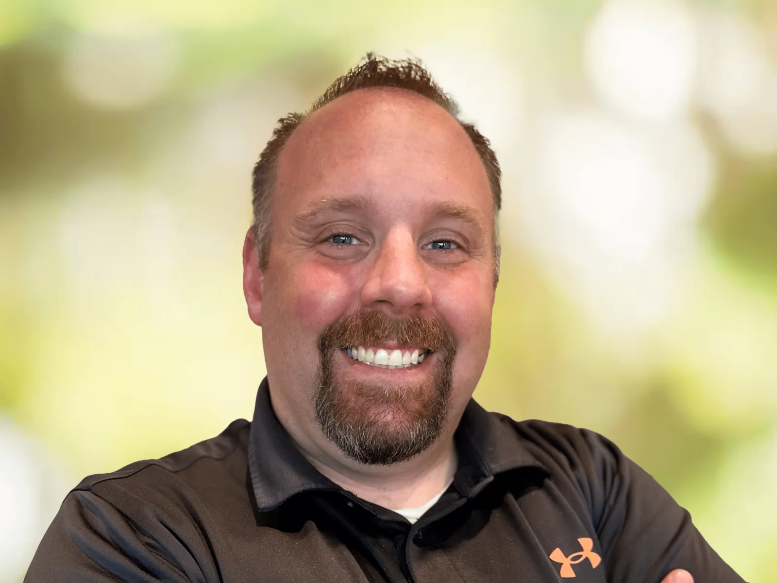 Smiling man in white and gray polo shirt with 100% chiropractic logo against a blurred green background.