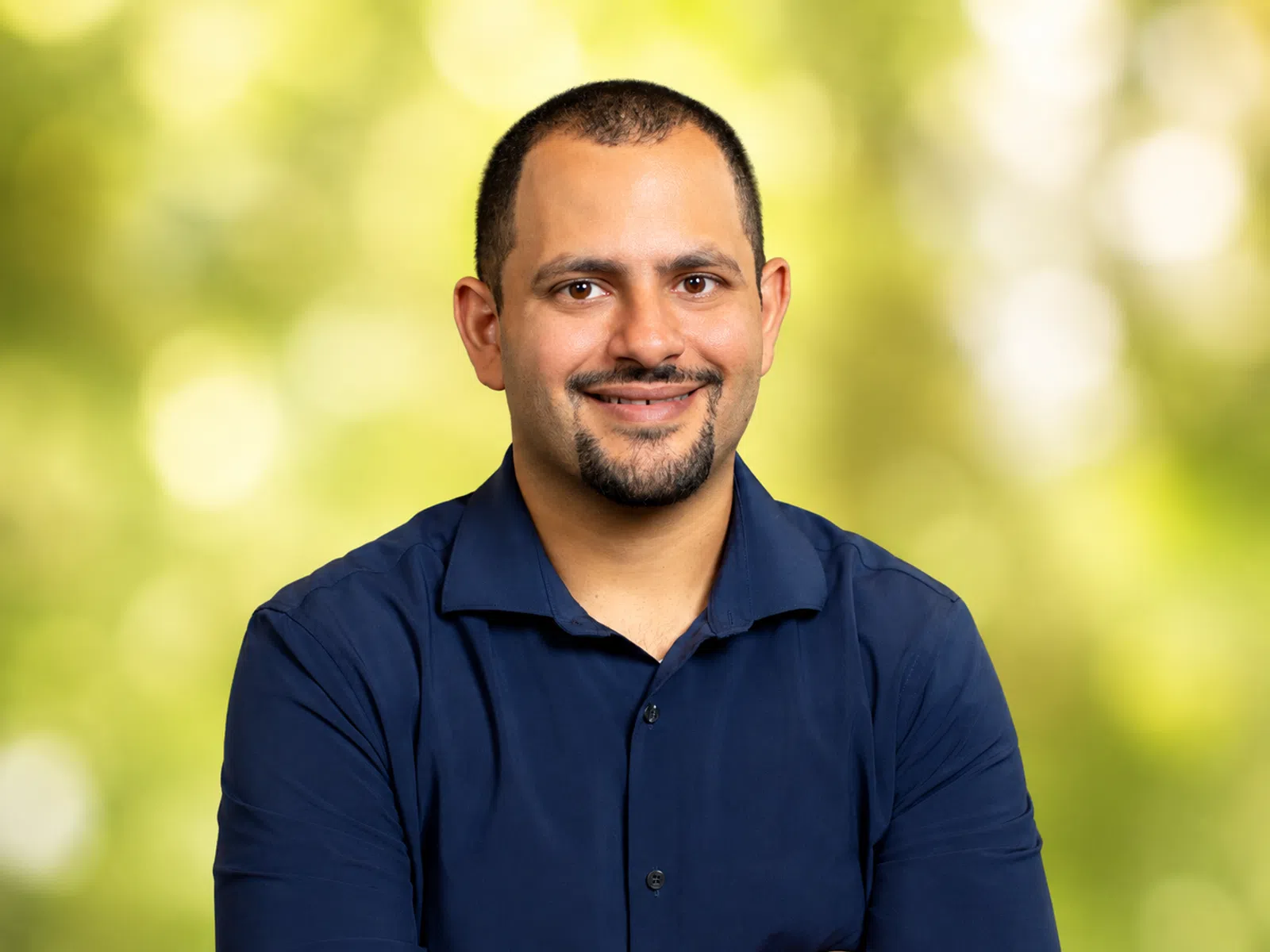 Smiling man with short hair and goatee wearing a navy blue shirt against a blurred green background.
