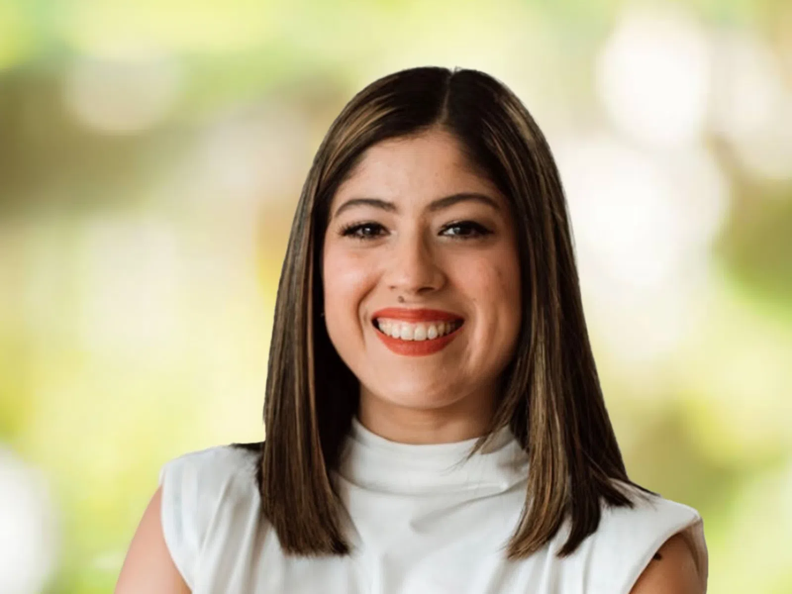 Smiling woman with straight brown hair wearing a white sleeveless top against a blurred green background