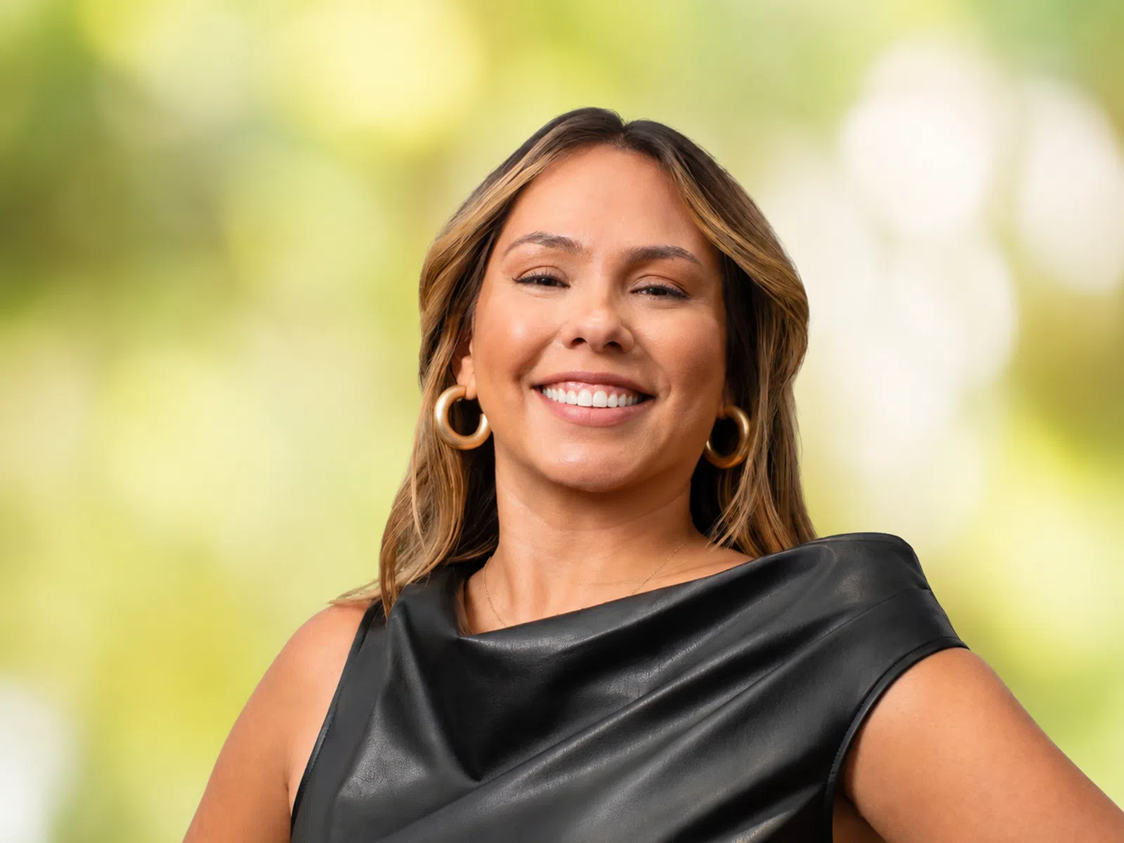 Confident woman smiling in black sleeveless leather top with gold hoop earrings against blurred green background