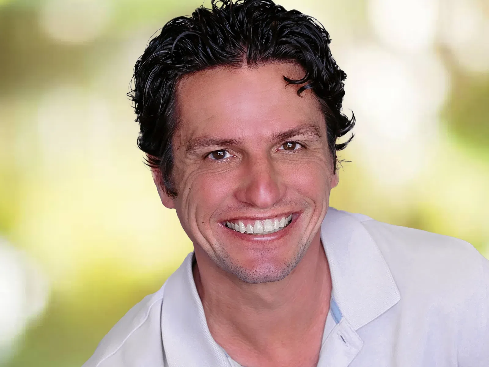 Close-up of a smiling man with curly black hair wearing a white polo shirt against a soft-focus green background.