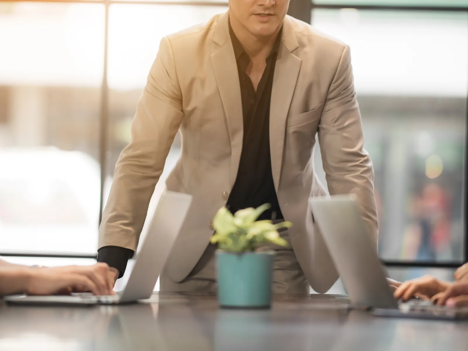 Man in beige suit leading a business meeting with laptops on a reflective table and natural light from windows.