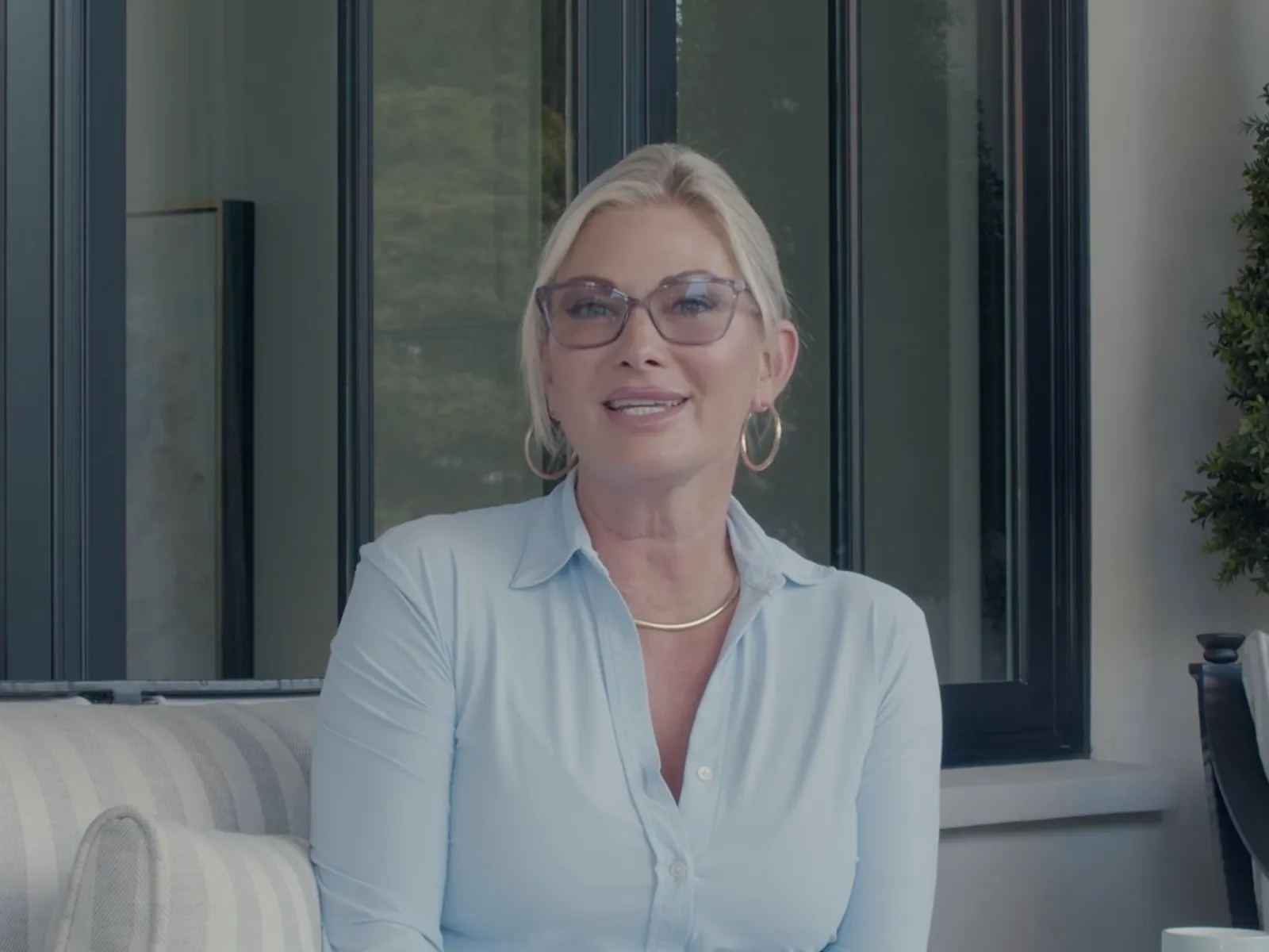 Blonde woman wearing glasses and light blue shirt sitting on a cushioned outdoor sofa near a window and plants.