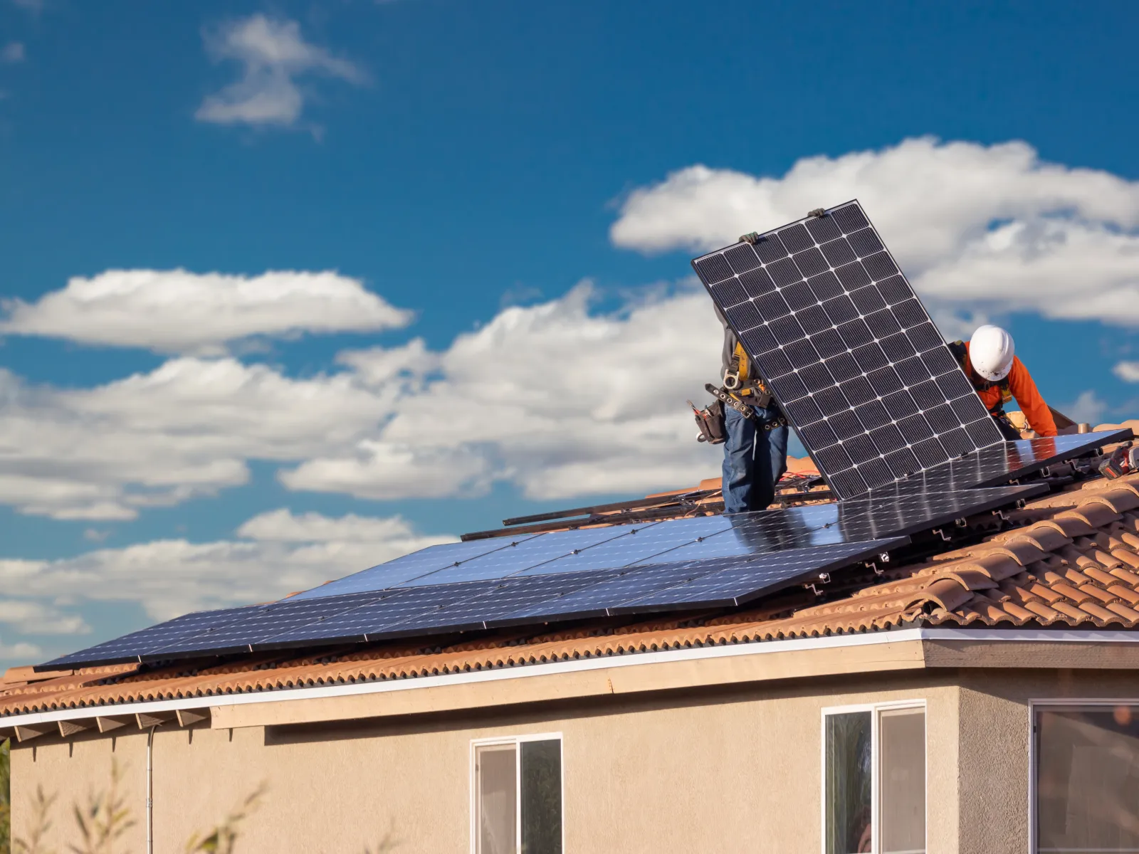 Workers installing solar panels on a tiled roof under a partly cloudy blue sky during daytime.