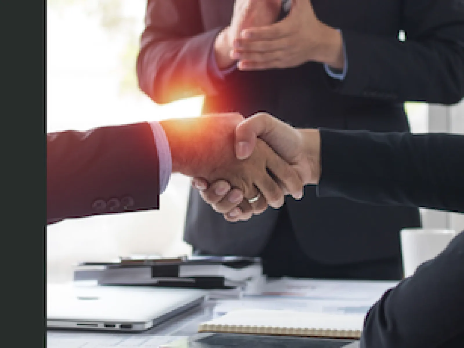 Business professionals shaking hands over a desk with a third person clapping in the background.