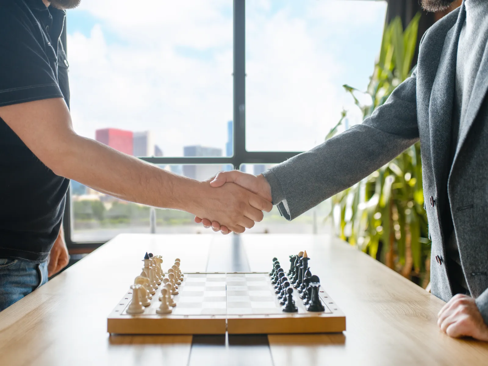 Two men shaking hands over a chessboard in a bright room with large window and plants in background