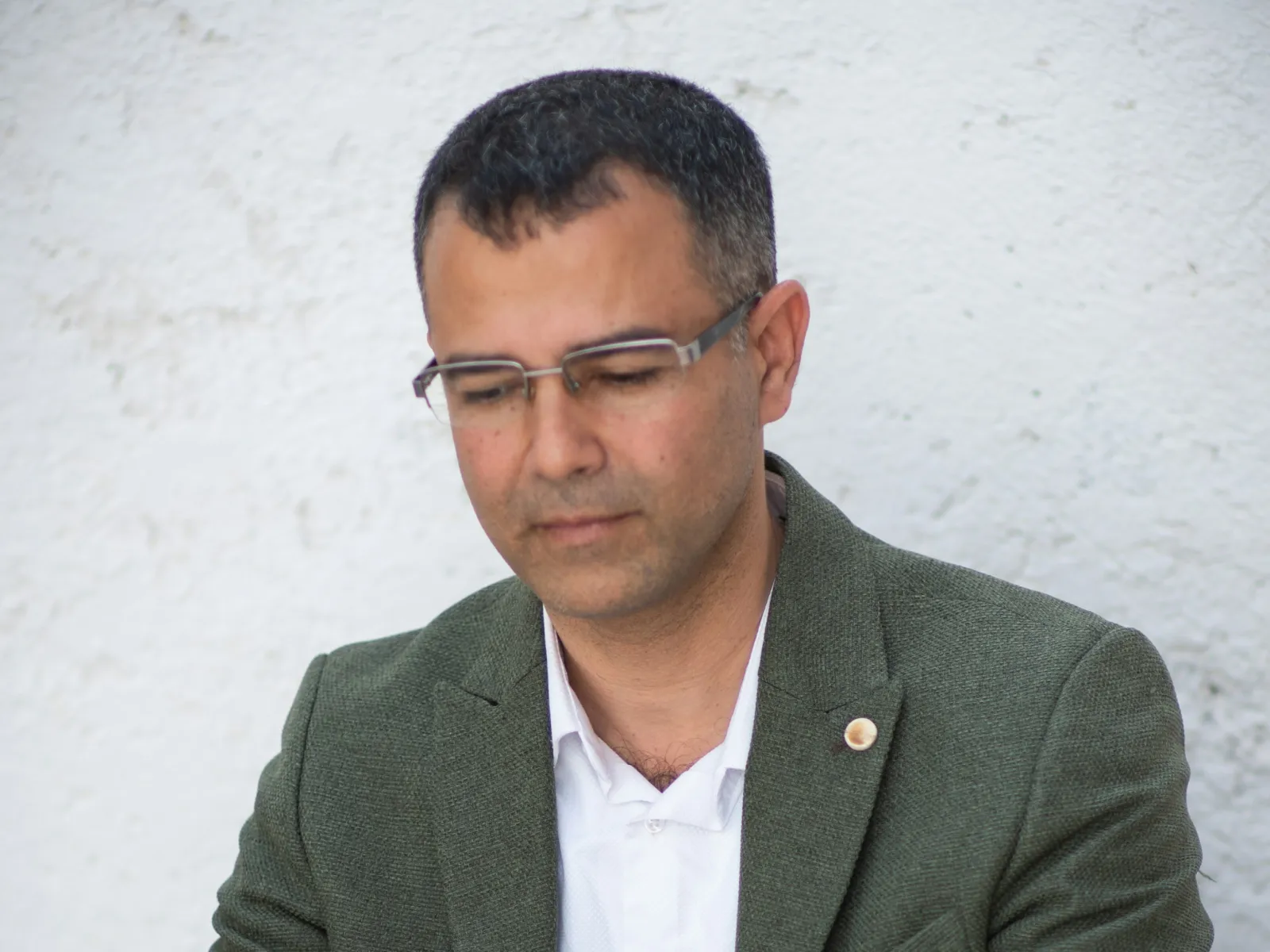 Man in green blazer working on a laptop at a wooden table with a book nearby against white textured wall.