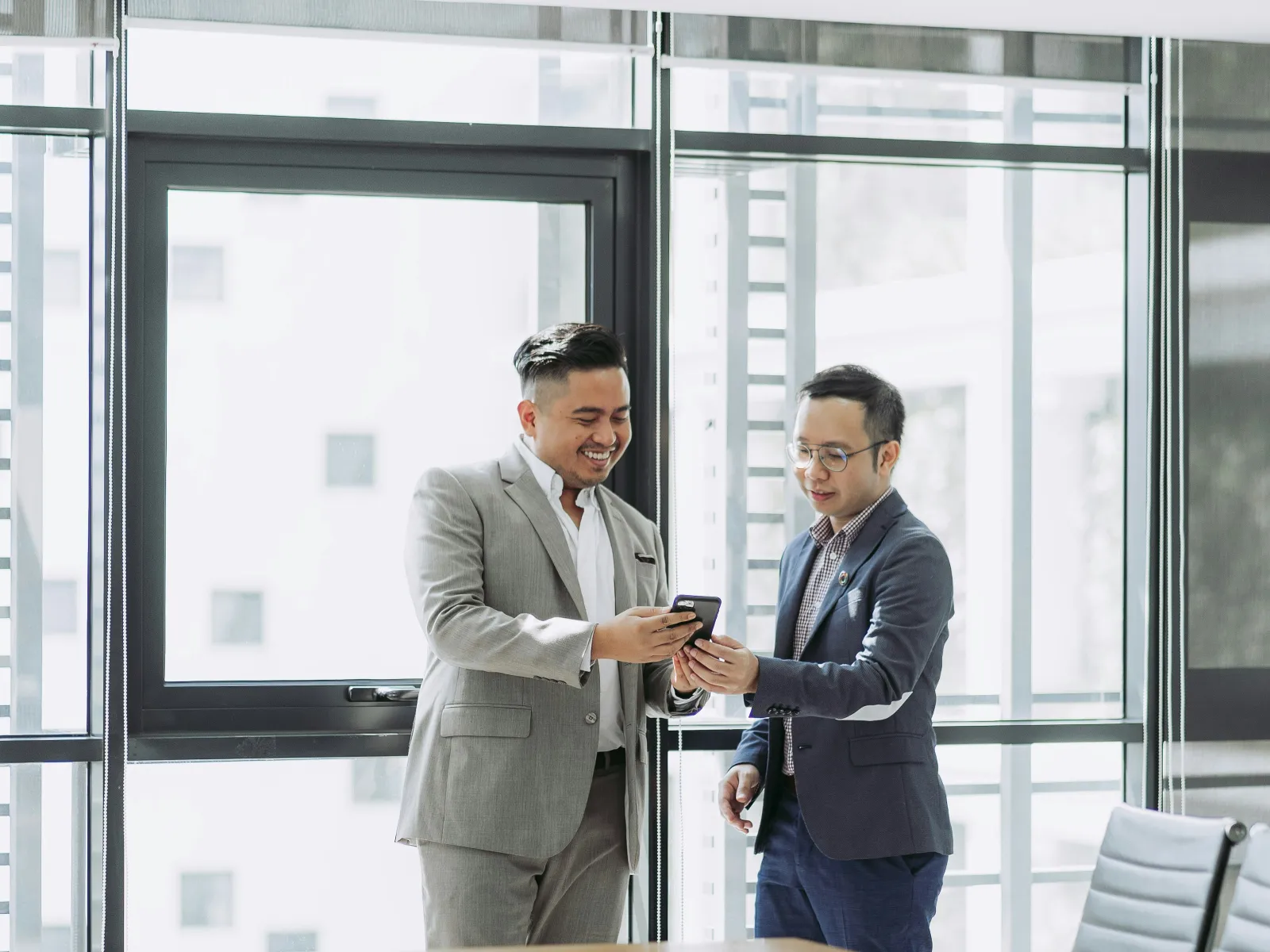 Two businessmen in suits sharing a smartphone in a modern conference room with large windows and chairs.