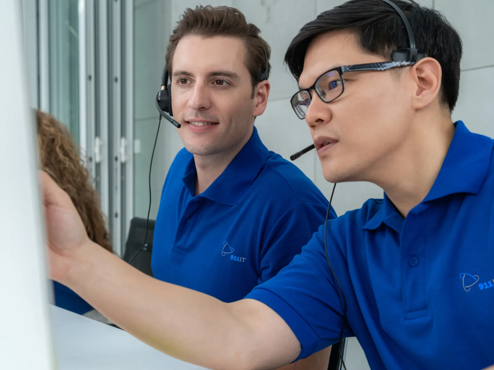 Two male IT support agents in blue shirts with headsets collaborating at a desk with a computer screen.