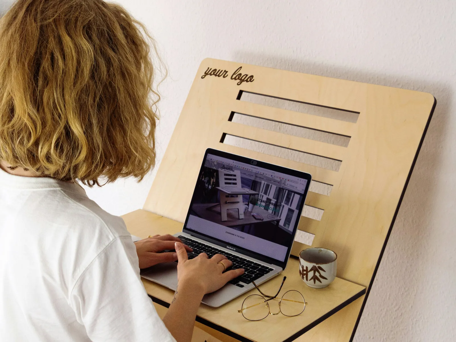 Woman using a standing wooden desk with laptop, coffee cup, and glasses, in a minimalist room.