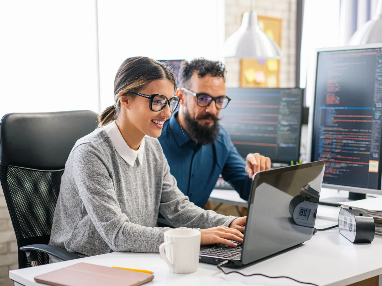 Two programmers collaborating on code at a workstation with laptops and monitors in a modern office.