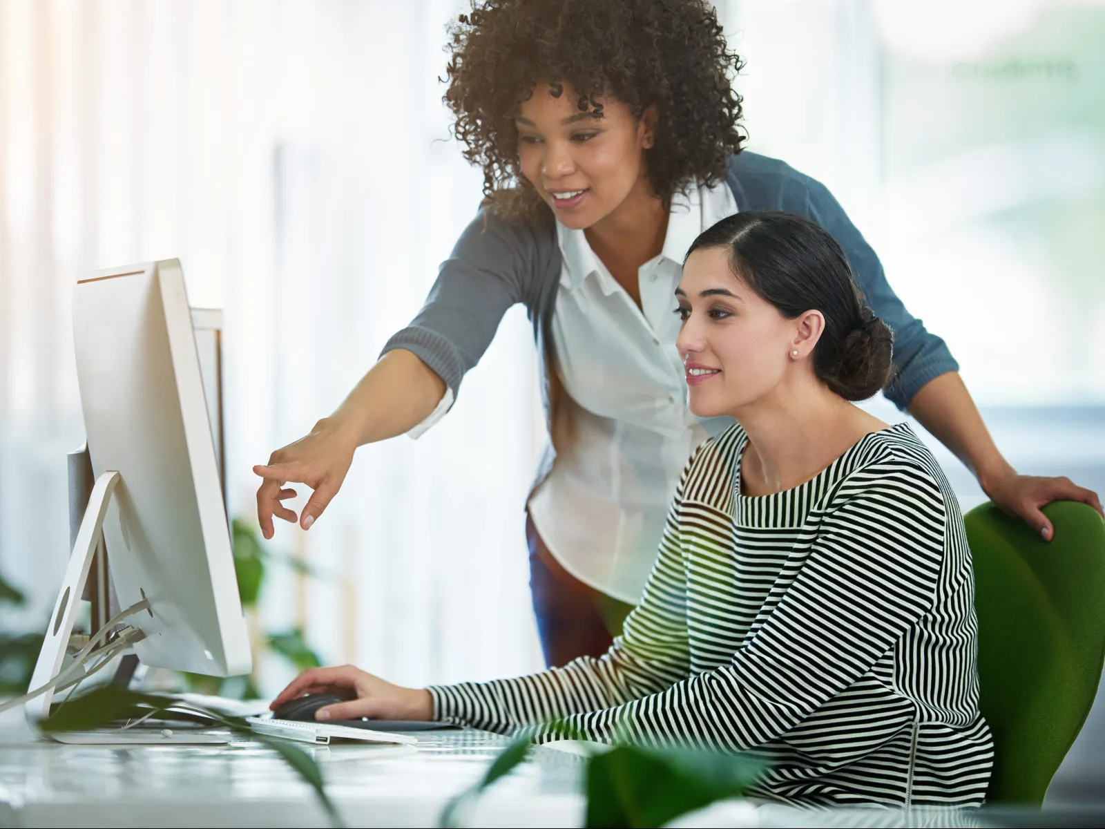 Two women collaborating at a computer, one seated and the other pointing at the monitor in a bright office environment