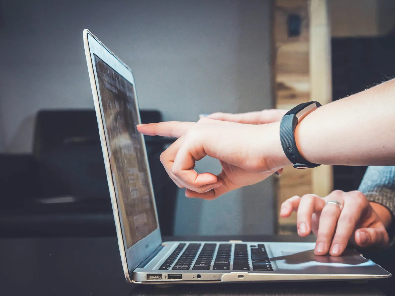 Person pointing at laptop screen while another uses the keyboard in a casual office setting.