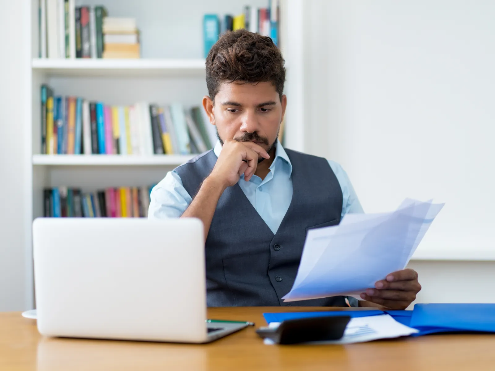 Focused businessman reviewing documents while working on laptop in a modern office with bookshelf background