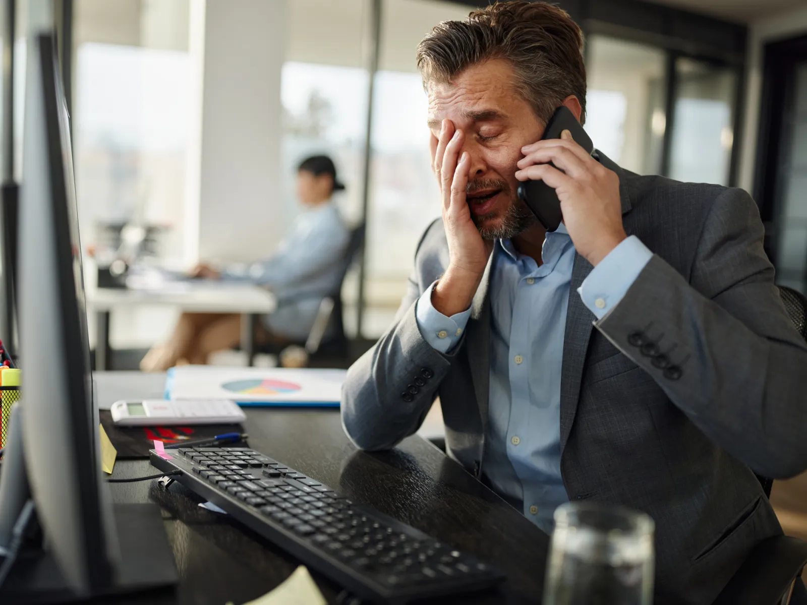 Stressed businessman in a suit talks on phone while covering his face at his office desk.