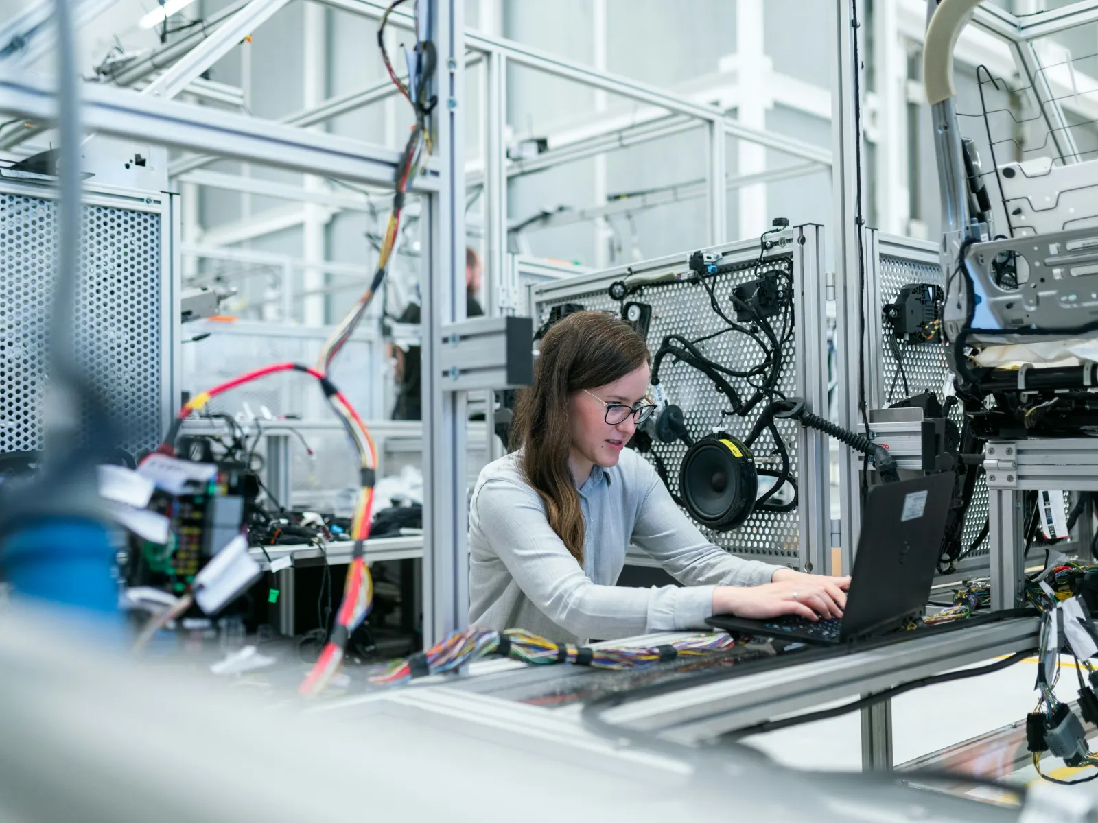 Young female engineer works on laptop in modern automotive manufacturing lab with car door and wiring systems.